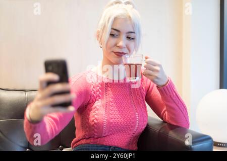 Eine junge Frau in einem rosafarbenen Pullover konzentriert sich intensiv auf ihr Smartphone und zeigt einen konzentrierten Ausdruck in einer gemütlichen Innenumgebung mit sanfter Beleuchtung Stockfoto