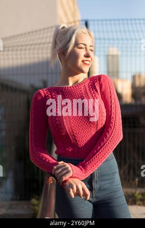 Eine fröhliche junge Frau in einem leuchtend roten Pullover lehnt sich draußen auf ein Geländer, lächelt und genießt das Sonnenlicht vor einem verschwommenen urbanen Hintergrund Stockfoto