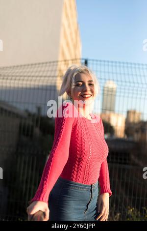 Eine fröhliche junge Frau in einem leuchtend roten Pullover lehnt sich draußen auf ein Geländer, lächelt und genießt das Sonnenlicht vor einem verschwommenen urbanen Hintergrund Stockfoto