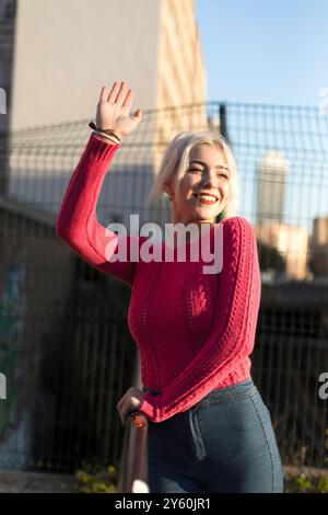 Eine fröhliche junge Frau in einem roten Pullover winkt mit einem hellen Lächeln und genießt die sonnige urbane Umgebung, ihr platinblondes Haar im Licht. Stockfoto