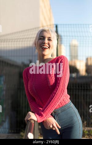 Eine fröhliche junge Frau in einem leuchtend roten Pullover lehnt sich draußen auf ein Geländer, lächelt und genießt das Sonnenlicht vor einem verschwommenen urbanen Hintergrund Stockfoto