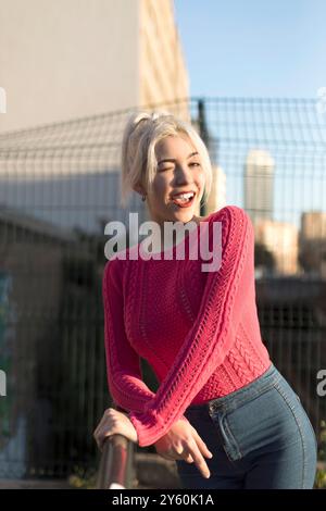 Eine fröhliche junge Frau in einem leuchtend roten Pullover lehnt sich draußen auf ein Geländer, lächelt und genießt das Sonnenlicht vor einem verschwommenen urbanen Hintergrund Stockfoto