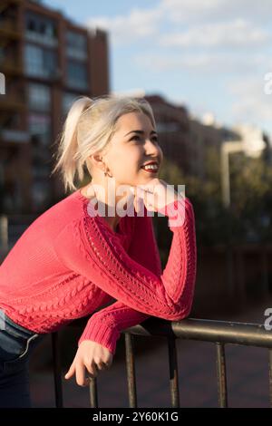 Eine junge Frau mit platinblonden Haaren lächelt, während sie sich auf ein Geländer stützt, in einen roten Pullover gekleidet und den Sonnenuntergang in einer urbanen Umgebung genießt Stockfoto