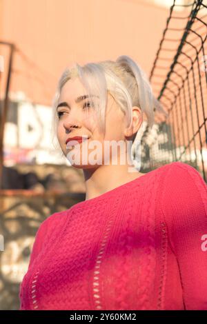 Eine fröhliche junge Frau in einem knallroten Pullover lächelt, während sie sich auf ein grünes Tor lehnt, mit urbanem Graffiti im Hintergrund, um einen sonnigen Tag zu genießen Stockfoto