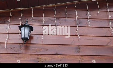 Wandleuchte in schwarzem Gehäuse mit weißem Milchglas und heller Girlande, die an einer hölzernen Keramikwand unter dem Dach des Hauses aus horizontalen Brettern hängt Stockfoto