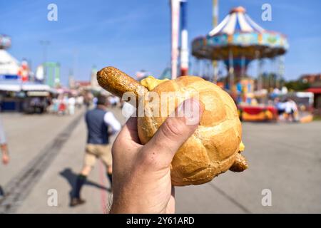 München, Bayern, Deutschland - 23. September 2024: Eine Hand hält eine vegane Bratwurst in einem mit Senf garnierten Brötchen auf dem Oktoberfest. Im Hintergrund sind verschwommene Eindrücke vom Festivalgelände und Menschenmassen im Hintergrund verschwommene Eindrücke vom Festivalgelände und Menschenmassen *** eine Hand hält eine vegane Bratwurst im Brötchen, die mit Senf garniert ist, auf dem Oktoberfest. Im Hintergrund sind unscharfe Eindrücke des Festgeländes und Menschenmengen Stockfoto