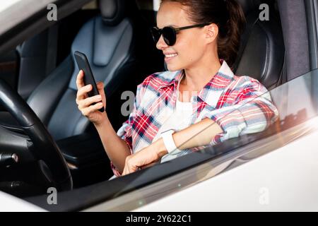 Online-Service für Autovermietung oder Carsharing. Junge Erwachsene Frau in lässiger Kleidung und Sonnenbrille, die in einem Auto sitzt und lächelt, während sie auf ihr Smartphone schaut. Stockfoto