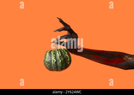 Schwarze Hexenhand mit Klauen und grünem Kürbis auf orangem Hintergrund. Halloween-Feier Stockfoto