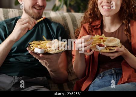 Zugeschnittene Aufnahme eines glücklichen jungen Paares, das Teller mit Pommes Frites und Hamburger hält, während es in Ruhe fernsehen oder Heimkino ansieht Stockfoto