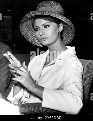 Elegante Frau, Filmstar Sophia Loren, cool in einem cremeweißen Anzug mit schlichten Linien und einem großen Cloche-Hut, erwischt in einer Lounge am Flughafen London, schlürft einen Drink und beobachtet die Welt wie sie vorbeizieht. Sophia wartete auf ihren Flug nach Rom. Sie war in diesem Land und machte die Millionärin mit Peter Sellers. Juli 1960 Stockfoto