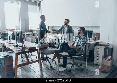 Willkommen an Bord! Hübsche junge Männer in lässiger Kleidung, die Hände schütteln und lächeln, während ihre Kollegen sie anschauen und lächeln Stockfoto