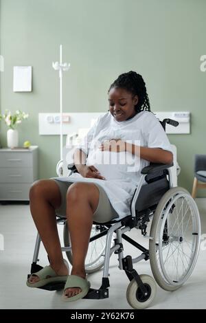 Afroamerikanerin, die im Rollstuhl sitzt und lächelt, während sie ihren Bauch in einem Krankenhauszimmer hält und freudige Vorfreude zeigt, leicht mit der Hand auf dem Bauch nach vorne gelehnt Stockfoto