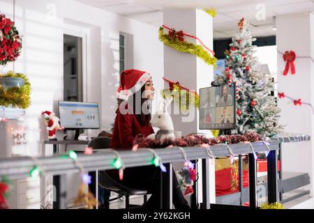 Erfahrener Mitarbeiter an festlich dekorierten Arbeitsplätzen mit weihnachtsmütze, chatten mit Kollegen bei Geschäftstelefonferenzen. Frau im Online-Video mit Kollegen während der Feiertage Stockfoto