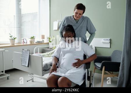 Krankenschwester, die Hilfe für schwangere Frau im Krankenhauszimmer mit medizinischen Geräten und komfortablen Einstellungen zur Verfügung stellt. Durch das Fenster einfallendes Tageslicht Stockfoto