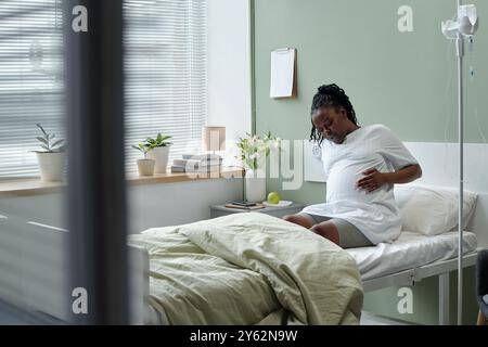 Schwangere Frau, die in Krankenhausbett nahe am Fenster mit IV-Stand in der Nähe und Klemmbrett an der Wand hängt Stockfoto