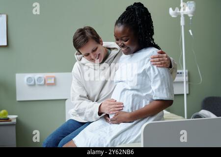 Kaukasischer Mann, der schwangere Schwarze Frau umschließt, die auf Krankenhausbett sitzt, lächelnd und bindend in medizinischem Umfeld Stockfoto