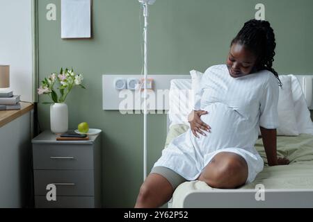 Lächelnde schwangere Frau, die ein weißes Kleid trägt, auf einem Krankenhausbett sitzt, ihren Bauch wiegt und nachdenklich nach unten blickt. Tisch mit Blumen und medizinischen Geräten im Hintergrund sichtbar Stockfoto