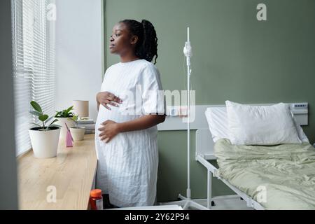 Schwangere Frau, die in der Nähe des Fensters im Krankenhauszimmer steht, während sie nach draußen blickt. Sie wirkt kontemplativ, ihre Hand sanft auf ihren Bauch gelegt, mit Krankenhausbett und Topfpflanzen in der Nähe Stockfoto