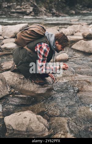Erfrischend mit kaltem Wasser. Hübscher junger moderner Mann, der Wasser aus dem Fluss trinkt, während er in den Bergen wandert Stockfoto