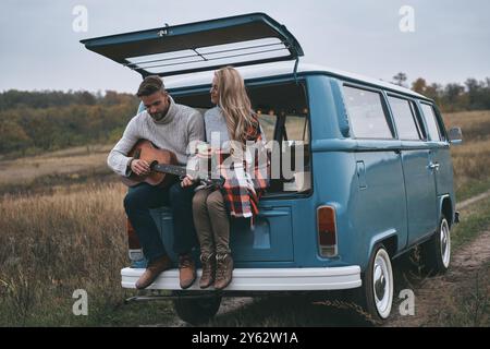 Romantisches Paar. Hübscher junger Mann, der Gitarre für seine schöne Freundin spielt, während er im Kofferraum eines blauen Minivans im Retro-Stil sitzt Stockfoto