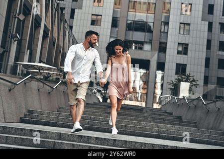 Die Liebe liegt in der Luft. Wunderschönes junges Paar, das Hände hält und lächelt, während man draußen die Treppe hinunter läuft Stockfoto