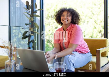 Lächelnde Frau, die einen Laptop benutzt, während sie auf einem gelben Stuhl sitzt, in einem hellen Zimmer, zu Hause Stockfoto