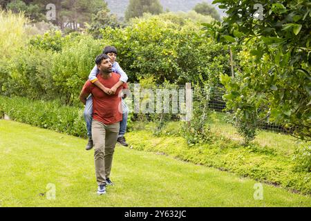 Indischer Vater, der den Sohn mit Huckepack reitet, während er in einem üppigen grünen Garten spaziert, Kopierraum Stockfoto