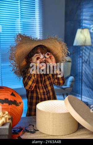 Süßer Junge, der wie Vogelscheuche gekleidet ist, mit dekorativen Augäpfeln, festlicher Dekoration und Geschenkboxen drinnen in der Nacht. Halloween-Feier Stockfoto