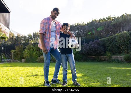 indischer Vater und Sohn halten Fußball und stehen zusammen auf Gras im Hinterhof Stockfoto
