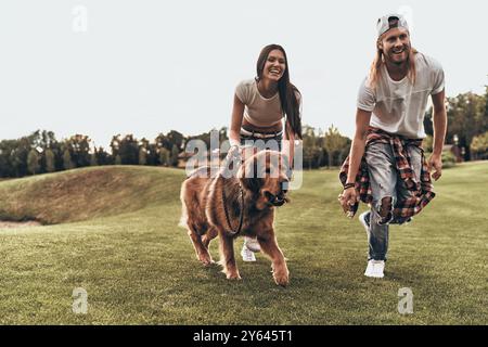 Perfekter, pelziger Freund. Die ganze Länge eines wunderschönen jungen Paares, das mit ihrem Hund spielt, während er im Park läuft Stockfoto