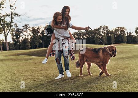 Wahre Liebe zum Ausdruck bringen. Die ganze Länge eines hübschen jungen Mannes, der eine junge attraktive Frau auf den Schultern trägt, während er mit seinem Hund im Freien geht Stockfoto