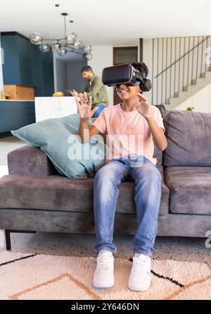 Mit dem VR-Headset sitzt ein indischer Junge auf der Couch, während der indische Vater im Hintergrund arbeitet Stockfoto