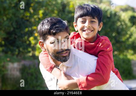 Ein indischer Vater reitet dem jungen Sohn mit Huckepack, beide lächeln draußen Stockfoto