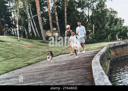 Guter Spaziergang. Die ganze Länge eines süßen kleinen Mädchens, das mit dem Hund läuft und lächelt, während ihre Eltern hinter sich her laufen Stockfoto