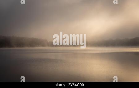 Eine ruhige nebelige Morgenlandschaft über einem ruhigen See mit sanftem Sonnenlicht, das durch den Nebel bricht. Stockfoto