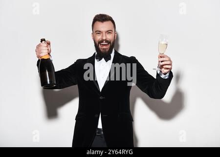 Lass die Party beginnen! Hübscher junger Mann im vollen Anzug, der eine Flasche Champagner hält und lächelt, während er vor grauem Hintergrund steht Stockfoto