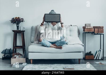 Geheimnisvolle Frau. Verspielte junge Frau in Freizeitkleidung und Strickmütze, die den kabellosen Lautsprecher vor dem Gesicht hält, während sie zu Hause auf dem Sofa sitzt Stockfoto