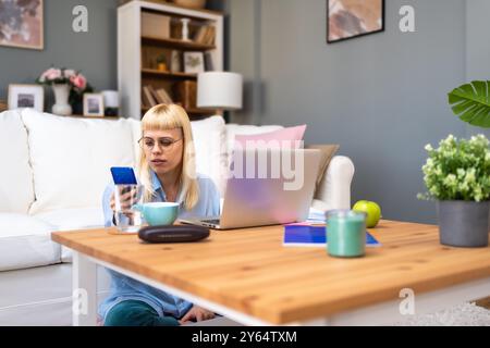 Junge Geschäftsfrau, die ihr Smartphone hält, während sie am Schreibtisch des Heimbüros mit einem Laptop arbeitet, und nutzt plattformübergreifende, responsive Design-Apps für die Projektanalyse Stockfoto