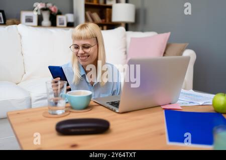 Junge Geschäftsfrau, die ihr Smartphone hält, während sie am Schreibtisch des Heimbüros mit einem Laptop arbeitet, und nutzt plattformübergreifende, responsive Design-Apps für die Projektanalyse Stockfoto
