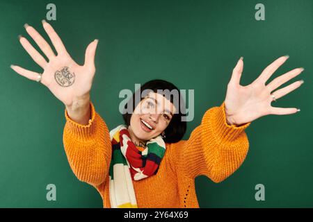 Eine junge Frau mit Tätowierungen lächelt hell, trägt einen gemütlichen Pullover und streckt ihre Hände spielerisch nach vorne. Stockfoto