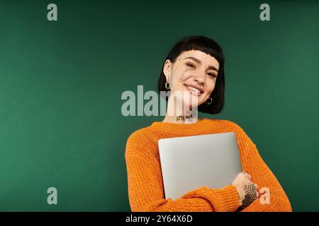 Eine junge Frau mit auffälligen Tätowierungen lächelt hell, während sie einen Laptop in einem pulsierenden Pullover umarmt. Stockfoto