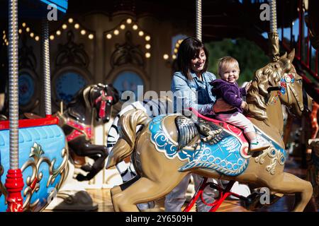 Eine Mutter und ihr lächelndes Kind reiten draußen auf einem bunten Karussell. Die Szene fängt Freude, Familienbindung und einen unbeschwerten Moment unter der Sonne ein. Stockfoto