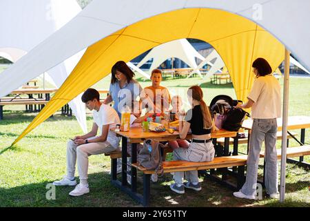 Eine Gruppe von Leuten versammelte sich um einen Tisch für ein Picknick unter einem gelben Baldachin. Die Umgebung ist im Freien und die Atmosphäre ist lässig und entspannt. Stockfoto