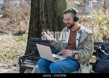 Junger Hipster-Expatriate freiberuflicher Arbeiter, der mit Laptop und kabellosen Kopfhörern im Park sitzt und Geschäftsvideos mit dem Kunden führt. Männlich IT engi Stockfoto
