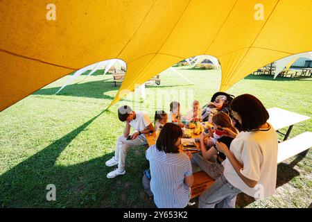 Eine Gruppe von Kindern und Familien versammelte sich zu einem Picknick unter einem großen orangen Baldachin an einem sonnigen Tag, bei Snacks und Freizeit im Freien. Stockfoto