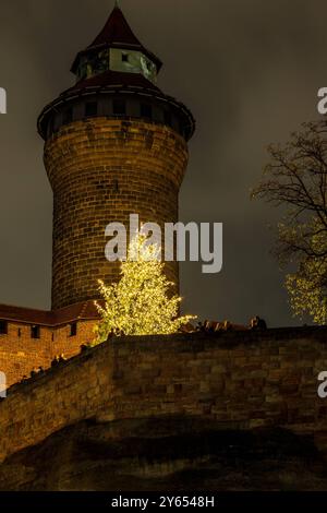 Nürnberger Christkindlesmarkt, 25.11.2022 der beleuchtete Turm der Kaiserburg in Nürnberg ragt in den dunklen Nachthimmel, während ein festlich geschmückter Weihnachtsbaum mit Lichterketten den Vordergrund ziert. Einige Menschen sind auf der Mauer vor dem Baum zu erkennen. Die Szene ist weihnachtlich und stimmungsvoll beleuchtet Nürnberg Bayern Deutschland *** Nürnberger Weihnachtsmarkt, 25 11 2022 in den dunklen Nachthimmel erhebt sich der beleuchtete Turm der Reichsburg Nürnberg, während ein festlich geschmückter Weihnachtsbaum mit Feenlichtern den Vordergrund ziert Stockfoto