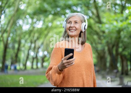 Reife Frau, die draußen im grünen Park Musik mit Kopfhörern und Smartphone hört. Ältere Frau genießt friedliche Momente, lächelt und zeigt Freude und Entspannung. Perfekt für Lifestyle-Konzepte. Stockfoto