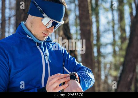 Sportlicher Mann in Wintersportbekleidung, der Smartwatch beim Workout im Wald verwendet, kaltes Wetter-Training. Er trägt eine reflektierende Sonnenbrille und ein Stirnband. Stockfoto