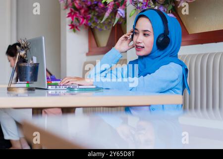 Schönes asiatisches Mädchen, das einen Schleier trägt und fröhlich vor dem Laptop lächelt und Musik hört, während er Kopfhörer in der Innenhalle hält. f Stockfoto