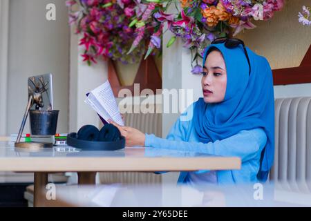 Geste einer schönen jungen asiatischen Frau, die einen eleganten Hijab trägt, der ein Buch liest und auf einem Sofa in einem Indoor Coffee Shop sitzt. Für Wissensinhalte: Lifest Stockfoto